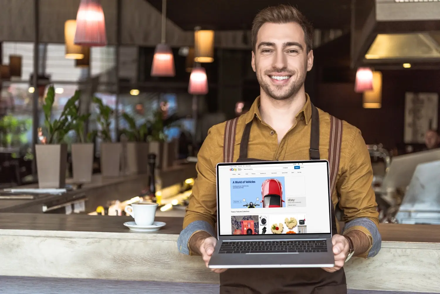 A person in a restaurant holds a laptop displaying an eBay webpage. They are standing behind a counter with a cup and saucer in front of them, perhaps discussing local SEO strategies to boost their online visibility.