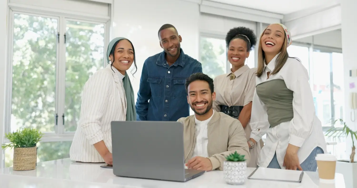 A group of five people smiling while standing and sitting around a table with an open laptop in an office setting, brainstorming ideas for website design for small businesses.